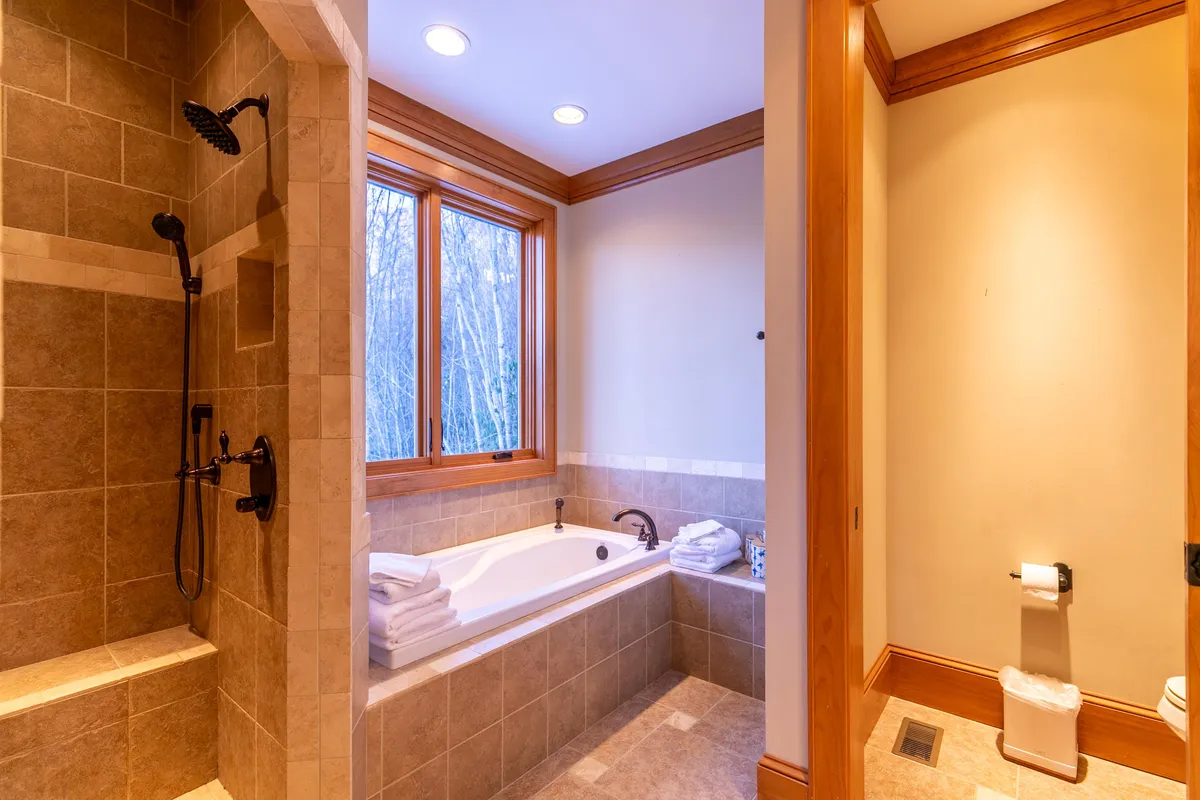 A spacious, spa-inspired bathroom with a large walk-in tile shower and a separate bathtub positioned by a window that offers views of the outdoors. The area is filled with soft natural light, evoking a sense of tranquility and rejuvenation.
