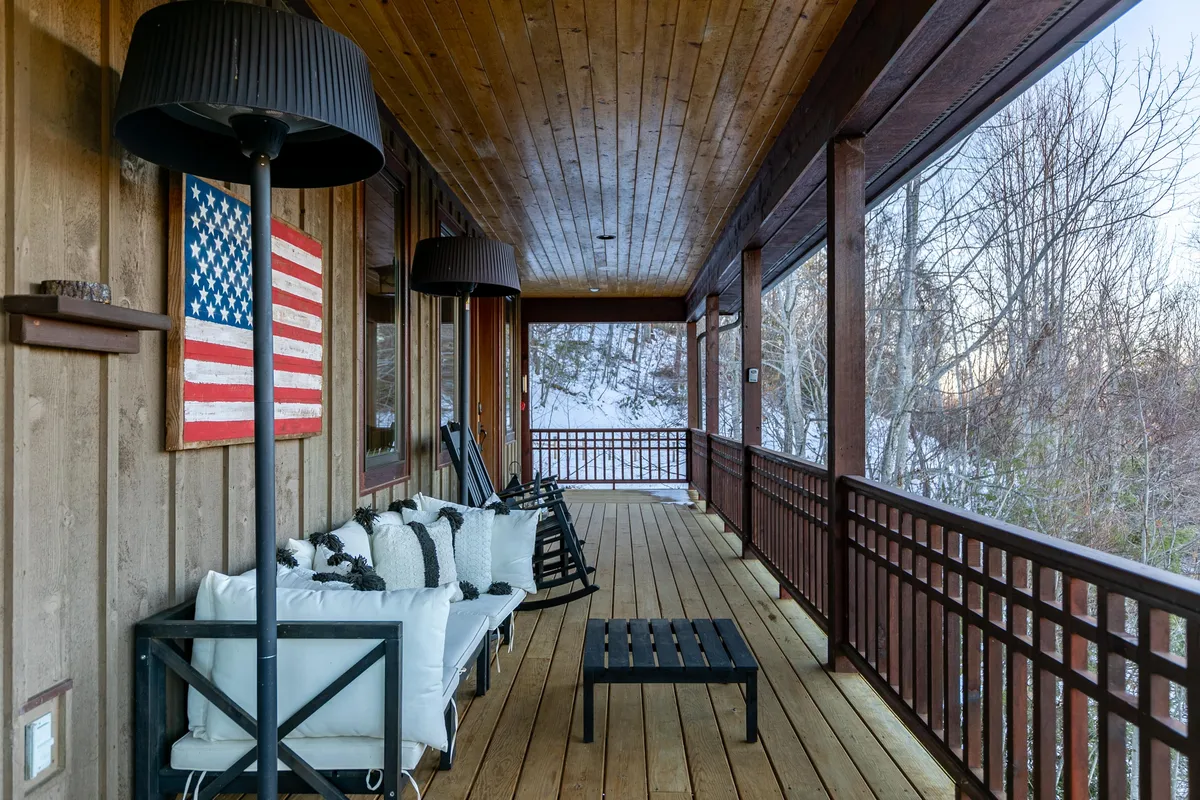 A cozy exterior porch with an American flag, black lamps, wooden railings, and a comfortable seating area overlooking the treetops. The setting conveys an inviting space for socializing, enjoying drinks or meals outdoors, and taking in the tranquil views.