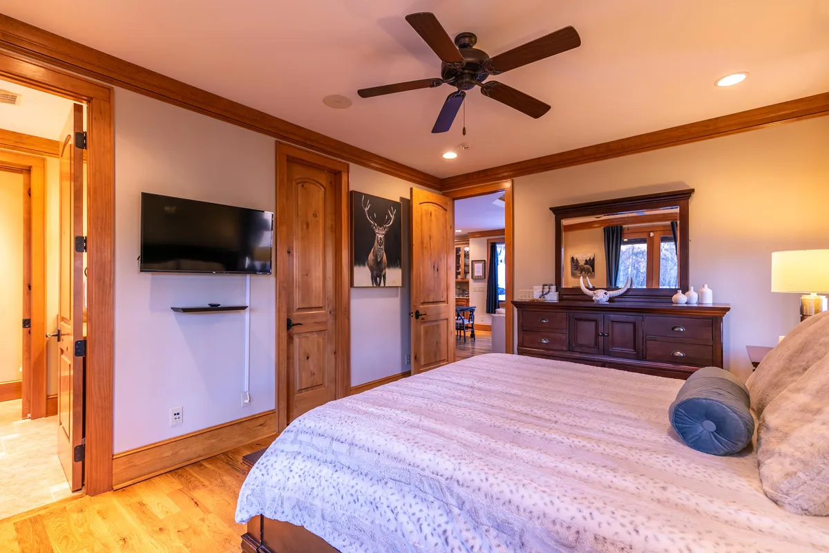 A warmly lit bedroom featuring a comfortable bed with plush bedding, a wall-mounted television, a dresser with a mirror, and charming deer artwork. Natural light filters through the window, creating an inviting and peaceful atmosphere ideal for rest and relaxation.