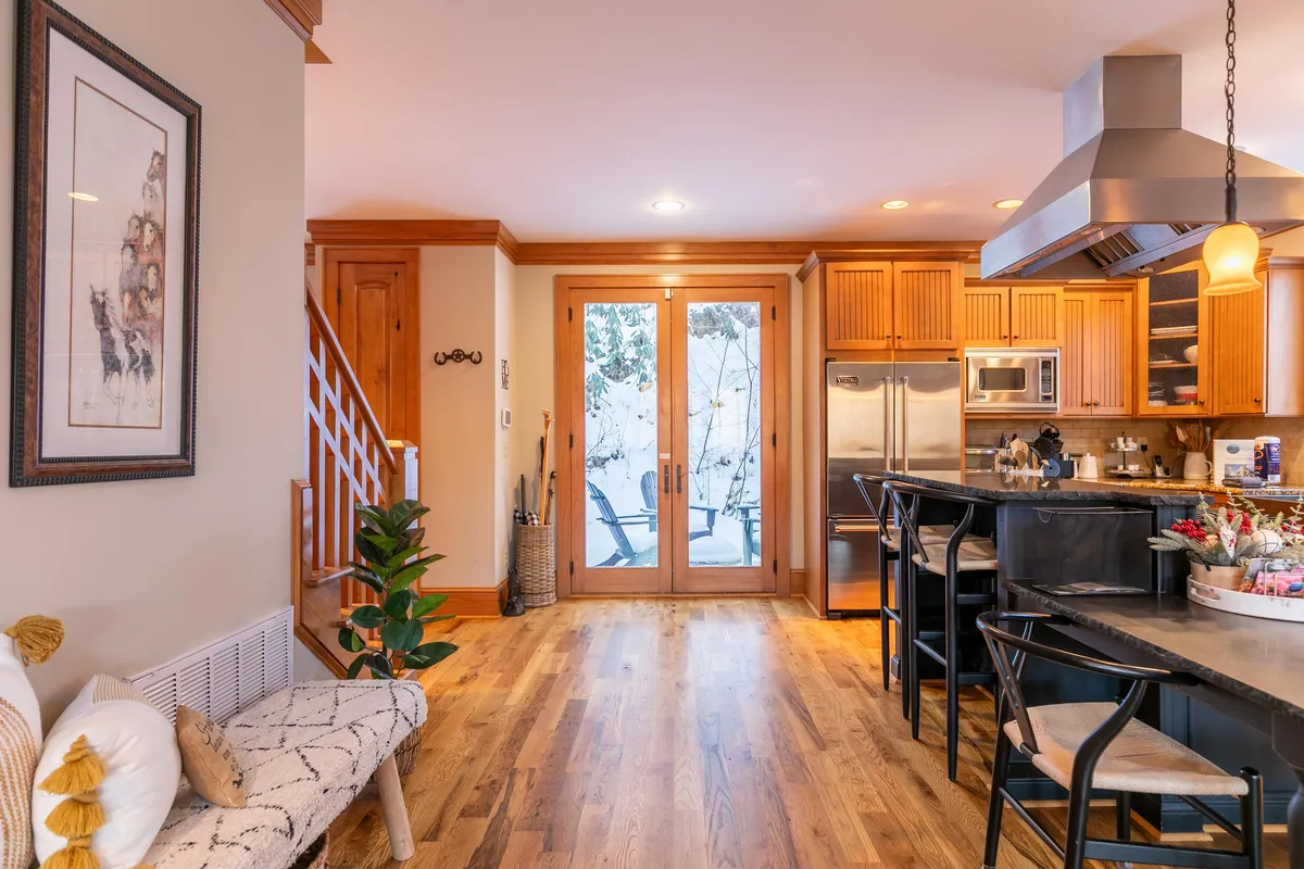A warm, inviting kitchen and dining space with double doors leading outside, wooden cabinetry, a kitchen island, and a staircase. The doors suggest easy access to outdoor areas, and the layout offers a sense of flow between indoor and outdoor living.