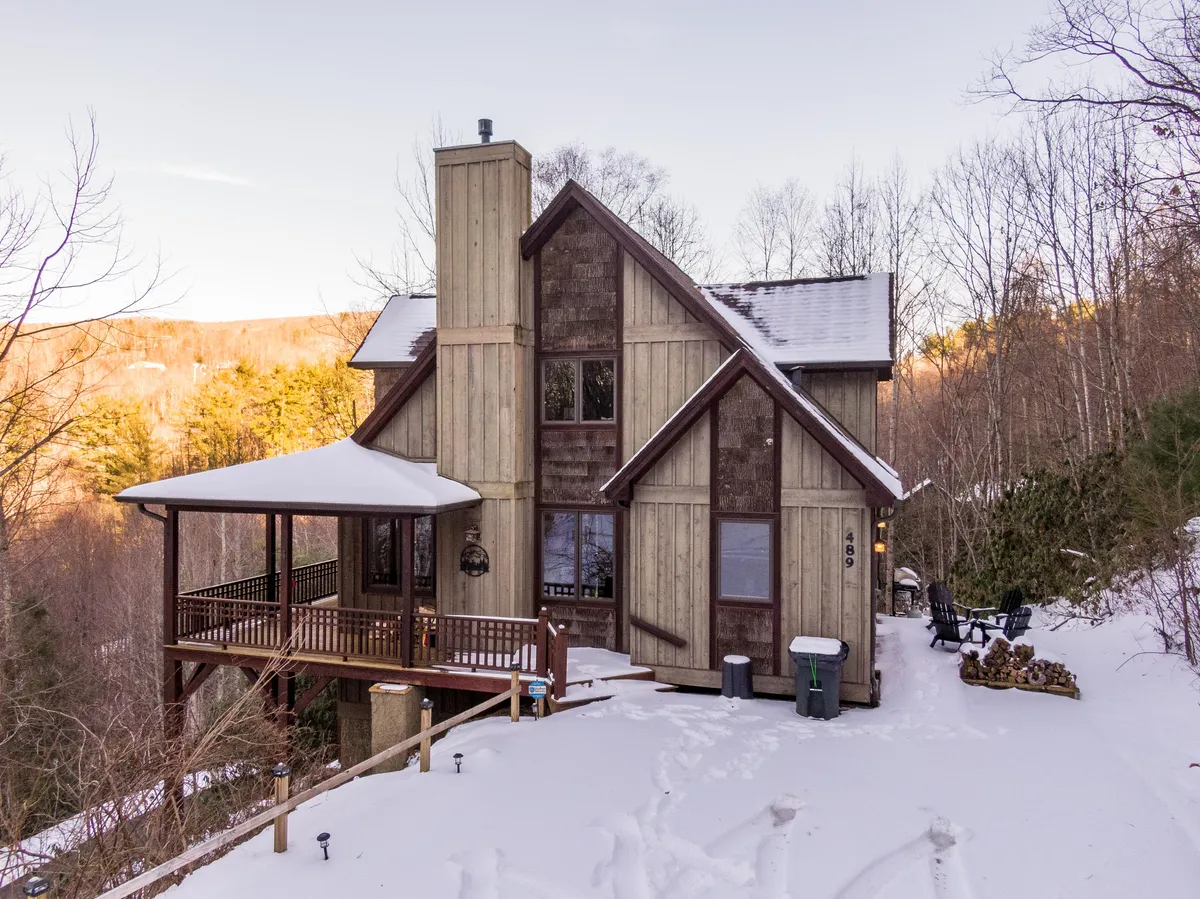 The deck with seating area overlooks snow-covered surroundings, with wooden siding and a stone chimney visible. The tranquil, wintry landscape and comfortable outdoor space invite guests to relax with a warm drink and take in the peaceful forest and mountain views.