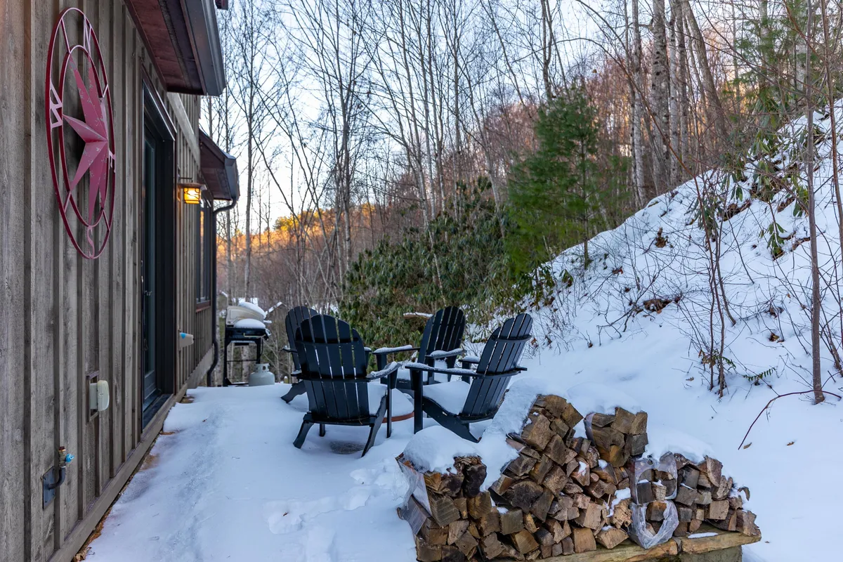 A tranquil exterior setting featuring Adirondack chairs gathered around a rustic firewood stack, a star decoration on the wall, and a snowy landscape. The scene evokes warmth and camaraderie, with inviting seating perfect for gathering friends to enjoy the mountain air and evening stories.