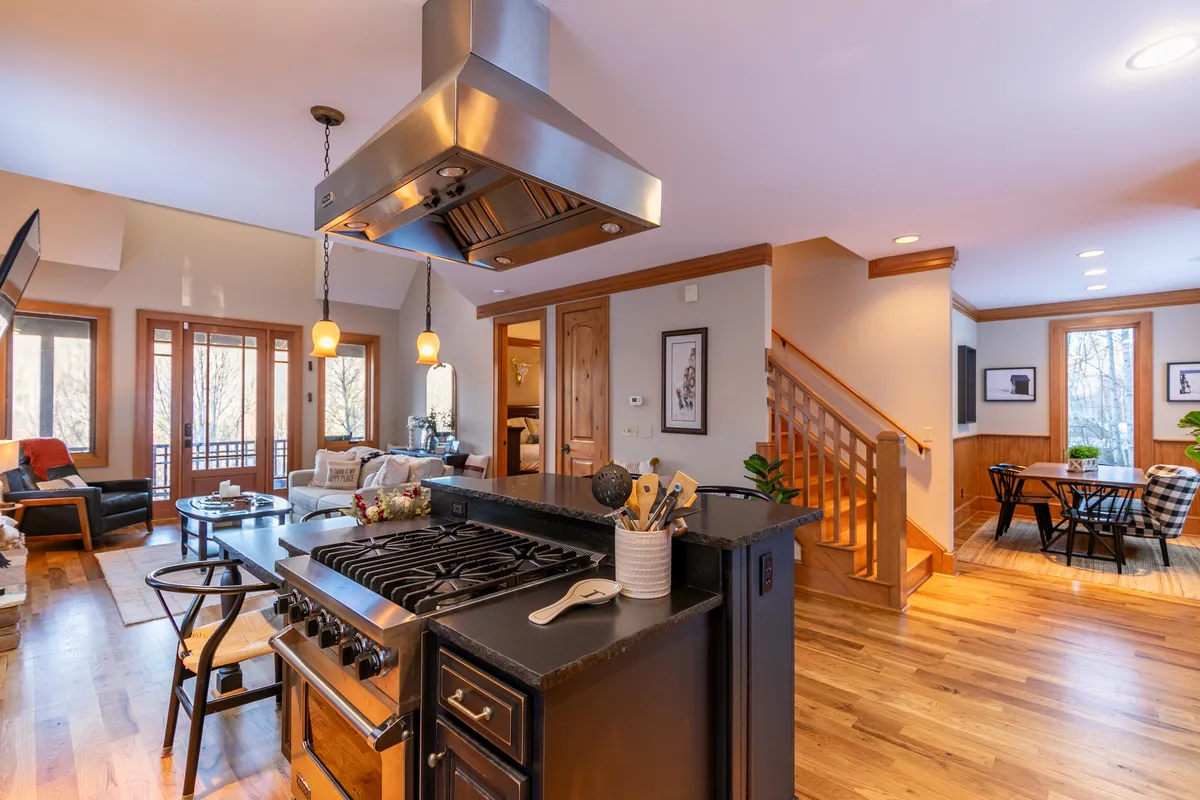 A wide interior view showing the kitchen island with stove, a living area with large windows, and a dining area with chairs, all in a single open-concept space. Warm lighting and wood accents enhance the inviting feel.