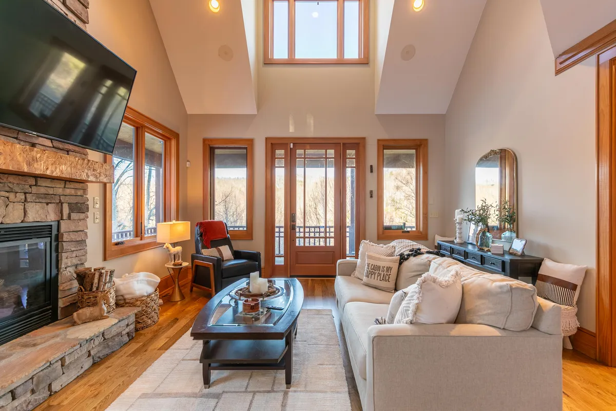 A cozy great room with a two-story stone fireplace as the centerpiece, flanked by large windows letting in natural light. Comfortable seating surrounds a coffee table, inviting guests to relax, entertain, or enjoy scenic views from indoors.