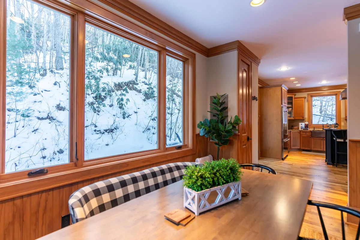 An inviting dining area with a large table, surrounded by windows that showcase views of the snowy outdoors. The space is warmed by natural light and decorated with simple, natural elements, ideal for sharing meals or relaxing while watching the seasons change.