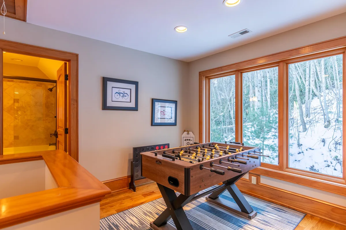 This image captures a cheerful loft space featuring a foosball table, large windows allowing ample natural light, framed artwork, and warm wooden accents. The setup suggests a lively environment for games and gatherings, with views that connect the loft to the outdoors.