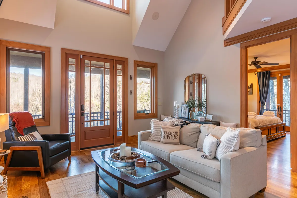 A cozy living room with a plush sofa, coffee table, large windows framing natural views, and an entrance door. The abundant sunlight and views create a sense of openness and connection to the wooded surroundings.