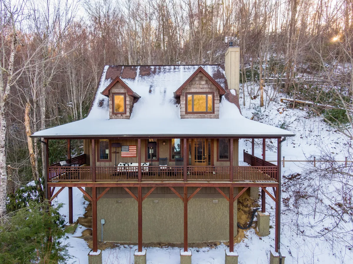 A picturesque snowy exterior of Headwaters Hideaway, featuring a welcoming front porch with an American flag, snow-covered roof, and large windows that hint at cozy indoor spaces. The porch and surrounding yard are ideal for guests—and their pups—to enjoy fresh air and scenic views.