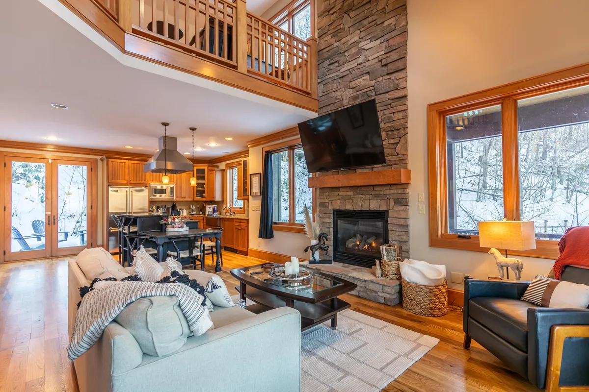 An open-plan space blending a warm seating area with a view of the kitchen island, fireplace, and expansive windows. The flow between living and kitchen spaces is evident, highlighting both relaxation and communal cooking.