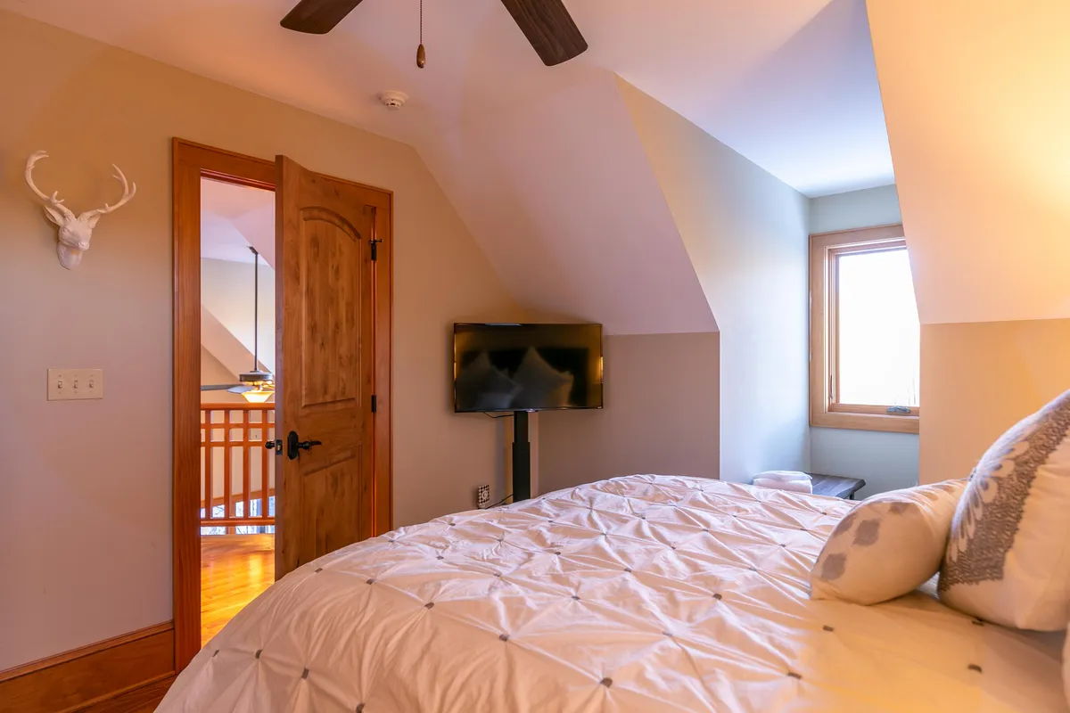 A cozy bedroom with a bed dressed in textured bedding, a TV mounted on the wall, a wooden door, and a window bringing in ample natural light. The ambiance is tranquil and inviting, perfect for restful nights and gentle mornings.